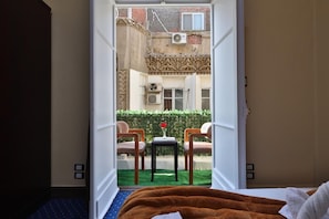 A hotel room with a bed, a brown blanket, a small table with a vase, and two chairs facing a garden with a hedge and a brick wall. - Cairo Paradise Hotel (Cairo)