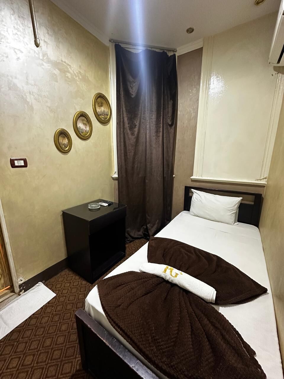 A hotel room with a bed, a bedside table, a wall-mounted light, a window with curtains, and a wall with three framed pictures.