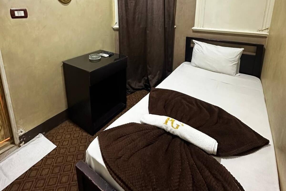 A hotel room with a bed, a bedside table, a wall-mounted light, a window with curtains, and a wall with three framed pictures.