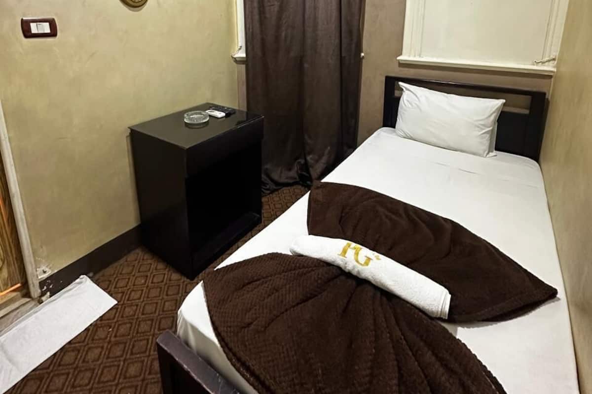 A hotel room with a bed, a bedside table, a wall-mounted light, a window with curtains, and a wall with three framed pictures.