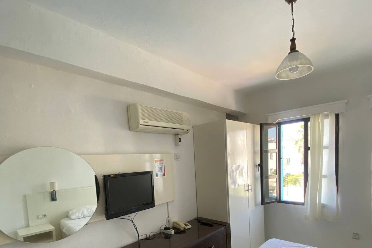 A hotel room with a bed, a desk with a flat-screen TV, a mirror, an air conditioning unit, and a window with curtains.