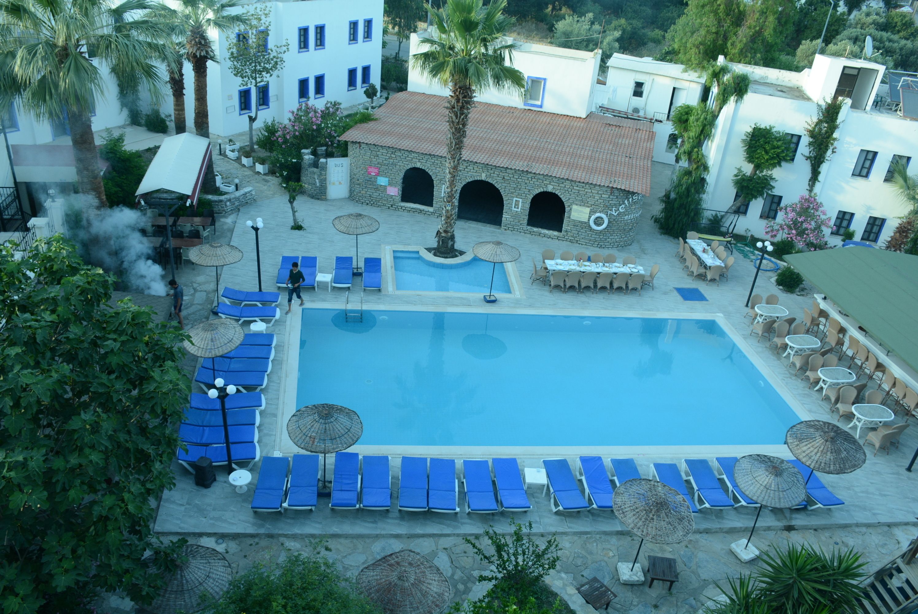 A pool area with blue lounge chairs and umbrellas, a stone building with arches, and palm trees.