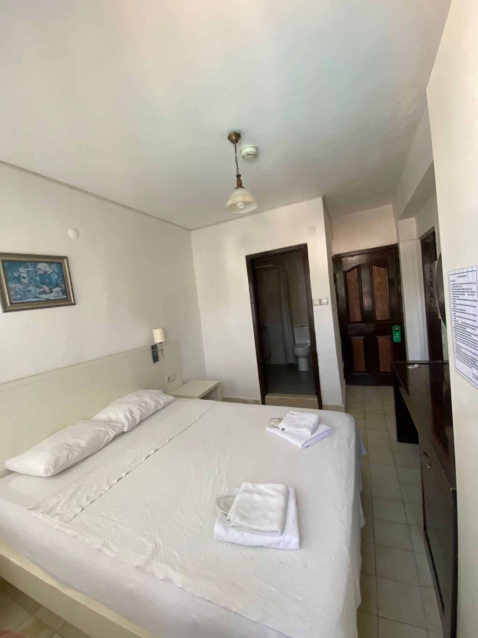 A hotel room with a large bed, a bedside table, a hanging light fixture, a framed picture on the wall, and a door leading to the bathroom.
