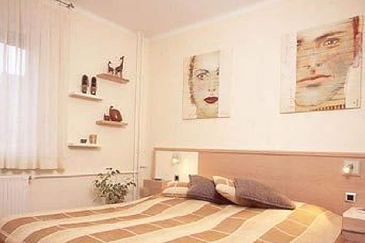 A bedroom with a large bed, a nightstand, a wall-mounted shelf with decorative items, and a window with curtains.
