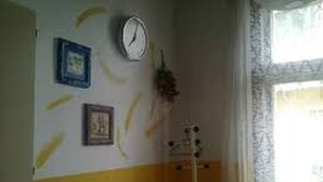 Una stanza con un orologio appeso al muro, una finestra con tende in pizzo e una chitarra appoggiata alla parete.