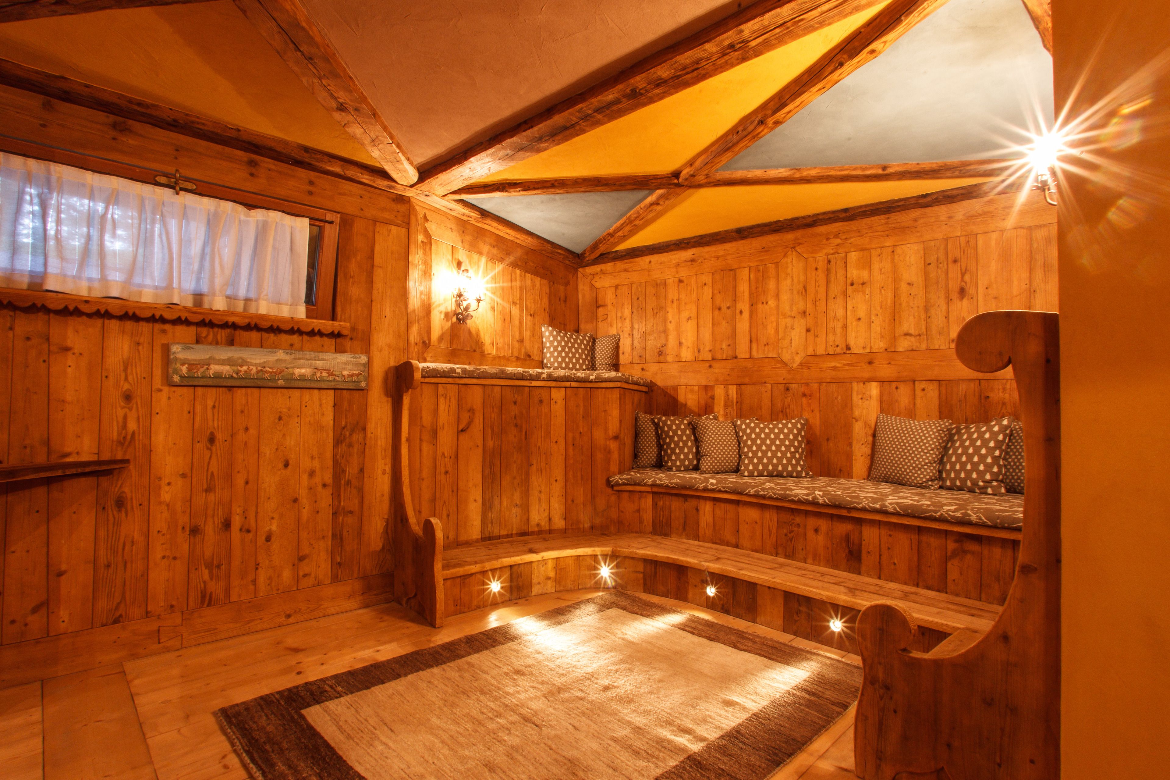 Interior de una cabaña de madera con un banco, un tapete y una lámpara fijada a la pared.