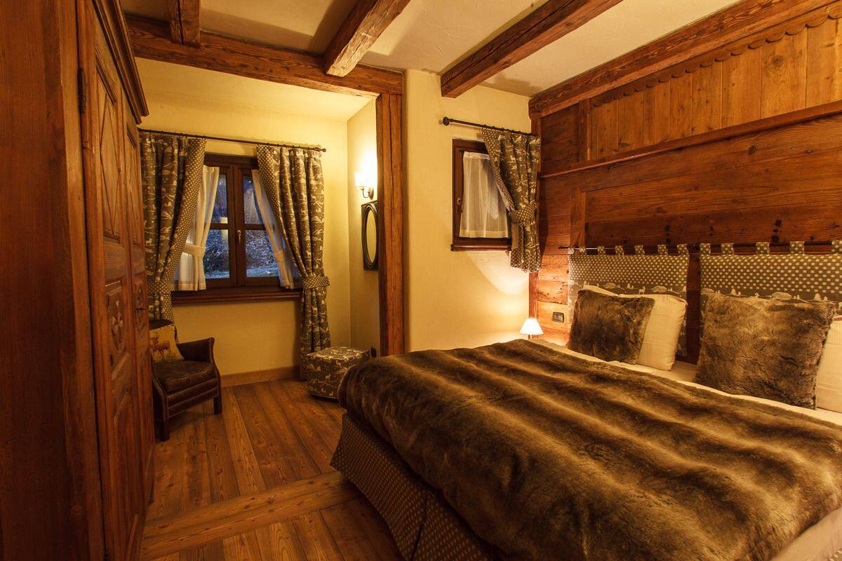Un dormitorio acogedor con cabecera de madera, una cama grande con manta oscura, una silla pequeña y una ventana con cortinas.