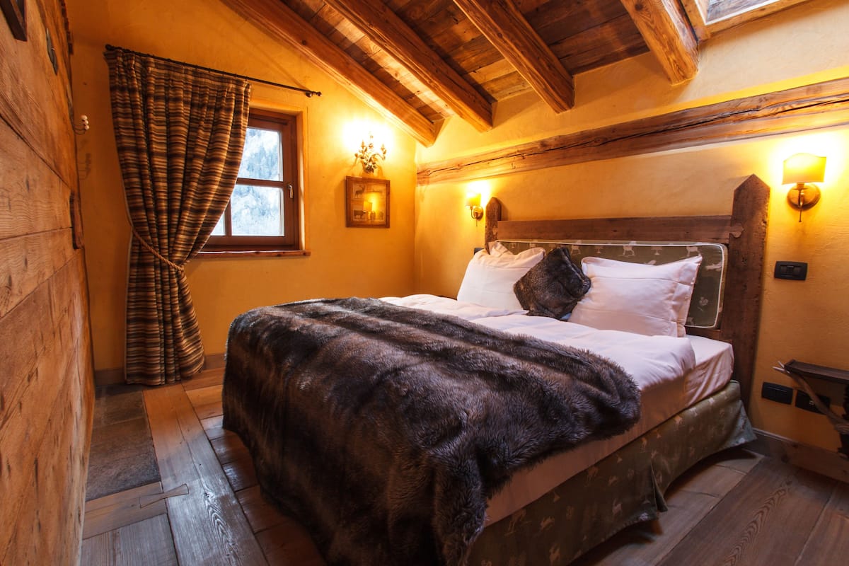 Una habitación acogedora con techo de madera, una cama con ropa de cama blanca, un plaid de piel oscura y una ventana con cortinas.