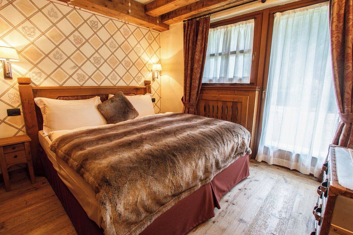 Una cama con estructura de madera y un cubrecama de piel, una mesita de noche con lámpara, y una ventana con cortinas.