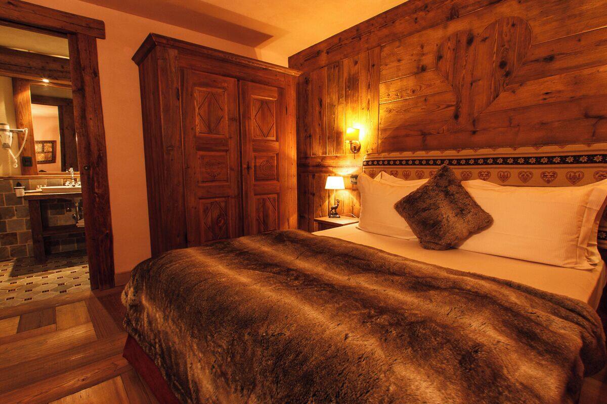 Una habitación con cabecera de madera, cama con una manta de piel, mesita de noche con lámpara y baño a la vista a través de una puerta abierta.