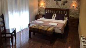 A bedroom with a stone wall, a bed with striped bedding, two bedside tables, a nightstand, a wooden chair, and a window with curtains. - Hotel Rustico Prado Da Viña (Fisterra)