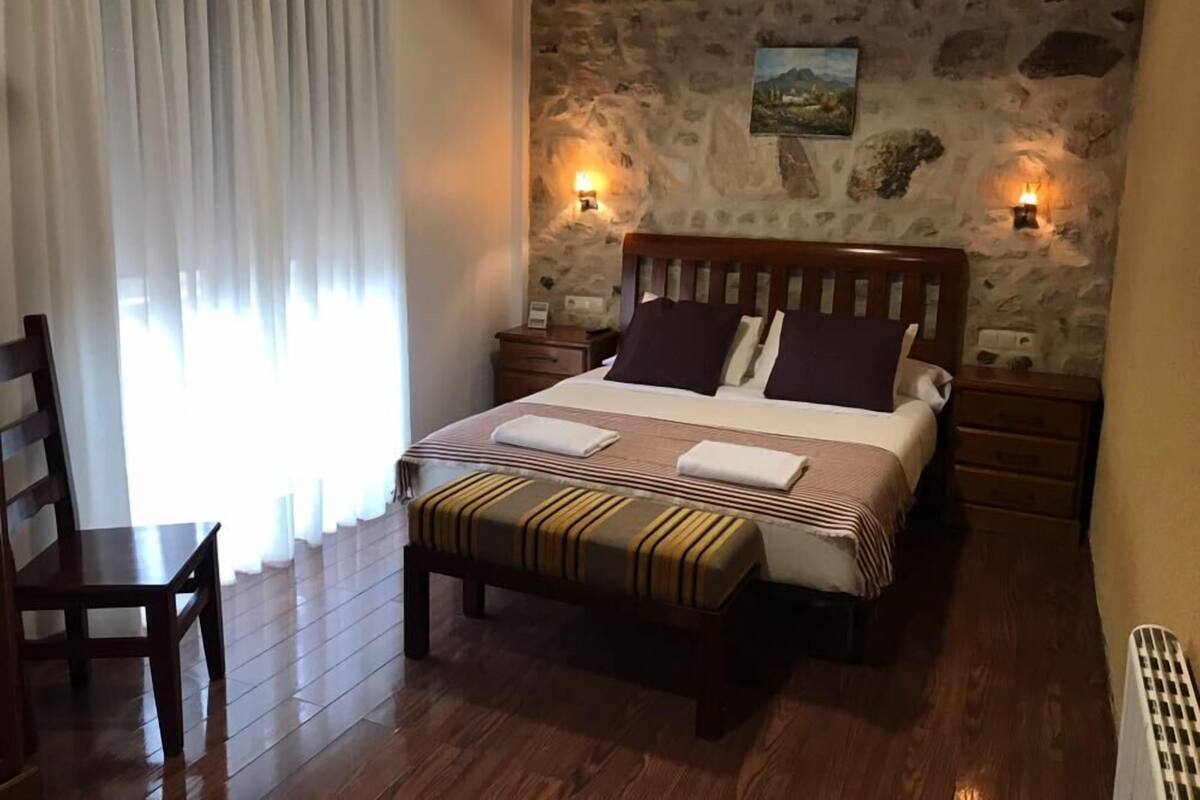 A bedroom with a stone wall, a bed with striped bedding, two bedside tables, a nightstand, a wooden chair, and a window with curtains.
