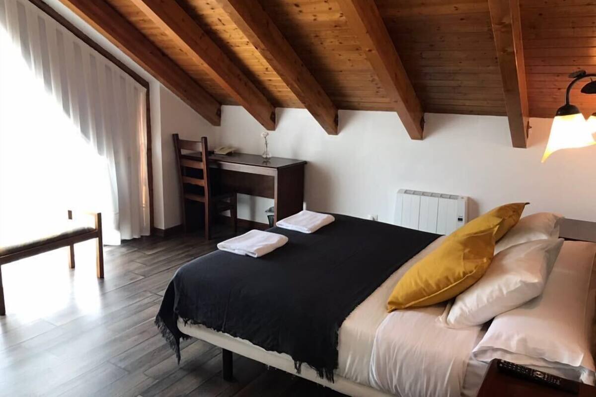 A bedroom with a wooden ceiling, a bed with white and yellow pillows, a bedside table, and a chair.