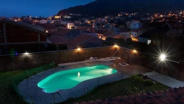 A swimming pool with a fountain, surrounded by a brick wall and a tiled deck, overlooking a residential area with houses and a hill in the background. - Hotel Rustico Prado Da Viña (Fisterra)