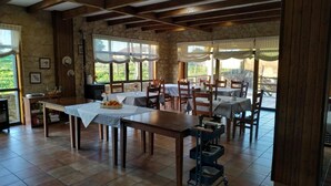 A dining area with stone walls, wooden beams, and large windows. Tables are set with checkered tablecloths and chairs. - Hotel Rustico Prado Da Viña (Fisterra)