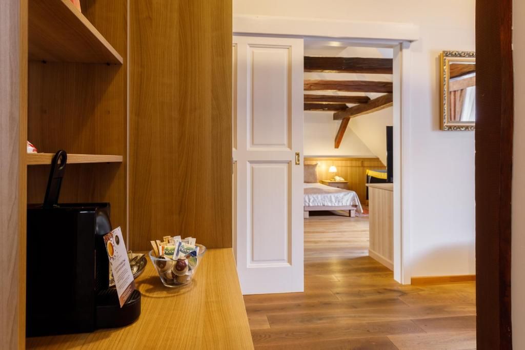 Habitación de hotel con piso de madera, cama con ropa blanca, una mesita con cafetera y un tazón de aperitivos, y techo de madera con vigas a la vista.