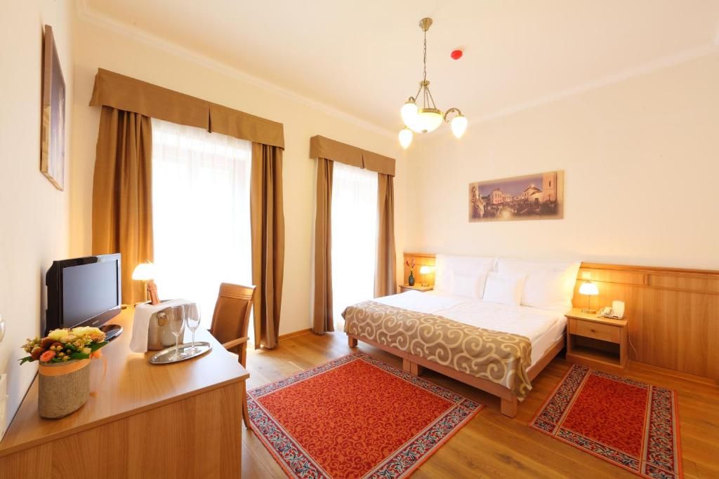 Quarto de hotel com cama, mesas de cabeceira, escrivaninha e um computador, uma cadeira e um tapete vermelho estampado.