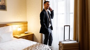 Una habitación de hotel con una cama, una mesita de noche con lámpara, una maleta y una ventana con cortinas.