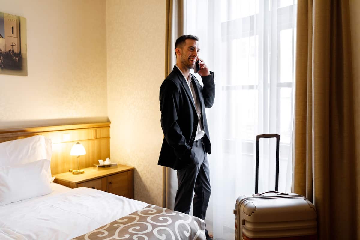 A hotel room with a bed, a bedside table with a lamp, a suitcase, and a window with curtains.