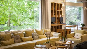 A cozy living room with a sofa, armchairs, and a coffee table, featuring a bookshelf and a large window with a view of greenery. - COCO-MAT Hotel Nafsika (Kifisia)