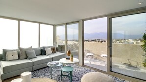 A modern living room with a grey sofa, a round table, and a view of the city and mountains through large windows. - COCO-MAT Hotel Nafsika (Kifisia)