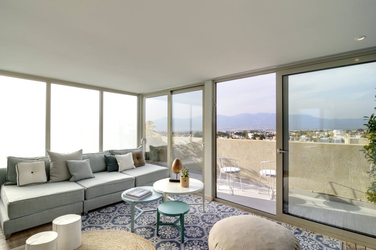 A modern living room with a grey sofa, a round table, and a view of the city and mountains through large windows.