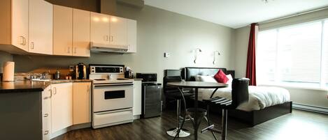 A modern kitchen with white cabinets, a stove, and a microwave. There is a dining area with a table and two chairs. The room has a large window with curtains.