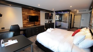 A modern hotel room with a large bed, a desk with a flat-screen TV, a wooden accent wall, and a separate bathroom with a glass shower. - Hotel Le Saint Germain (Rimouski)