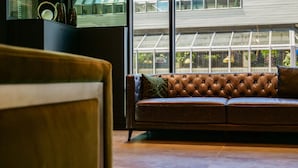 A brown leather sofa with a tufted backrest and a cushion, positioned in front of a large window with a view of a building and greenery. - Hotel Le Saint Germain (Rimouski)