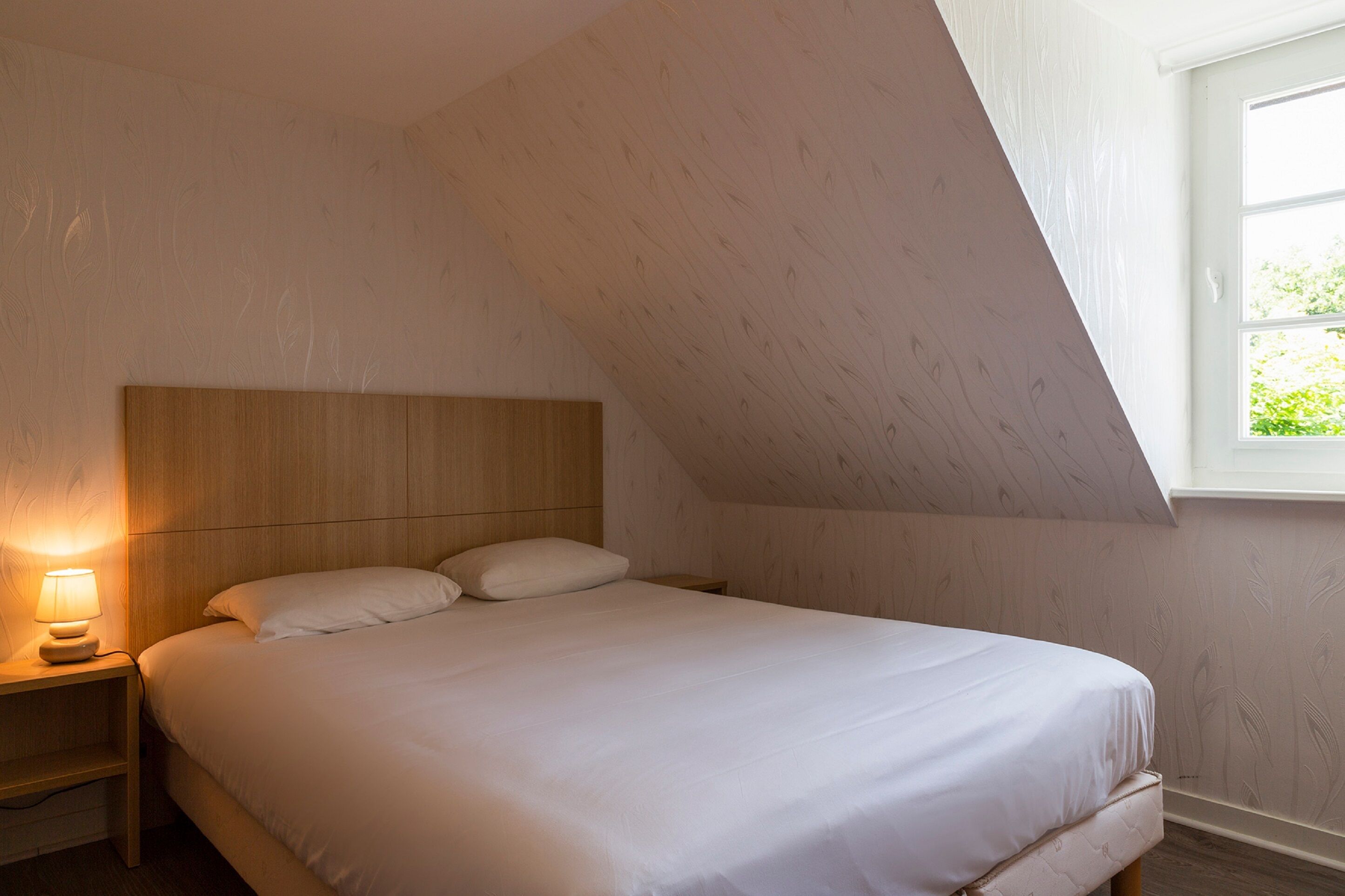 Une chambre à coucher avec une tête de lit en bois, une lampe de chevet et une fenêtre donnant sur un paysage verdoyant.