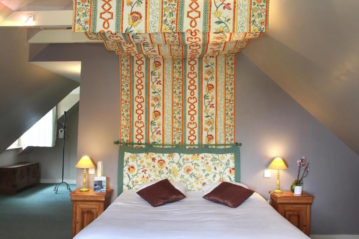 Un dormitorio con cama, dos mesitas de noche con lámparas, cabecera estampada y techo inclinado.