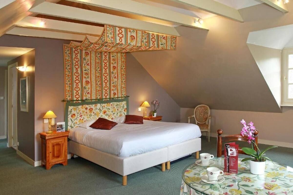 A bedroom with a bed, a nightstand with a lamp, a chair, a table with a teapot, and a window.