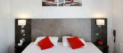 A hotel room with a large bed, two red pillows, a grey headboard, bedside lamps, and a wall-mounted artwork above the bed.