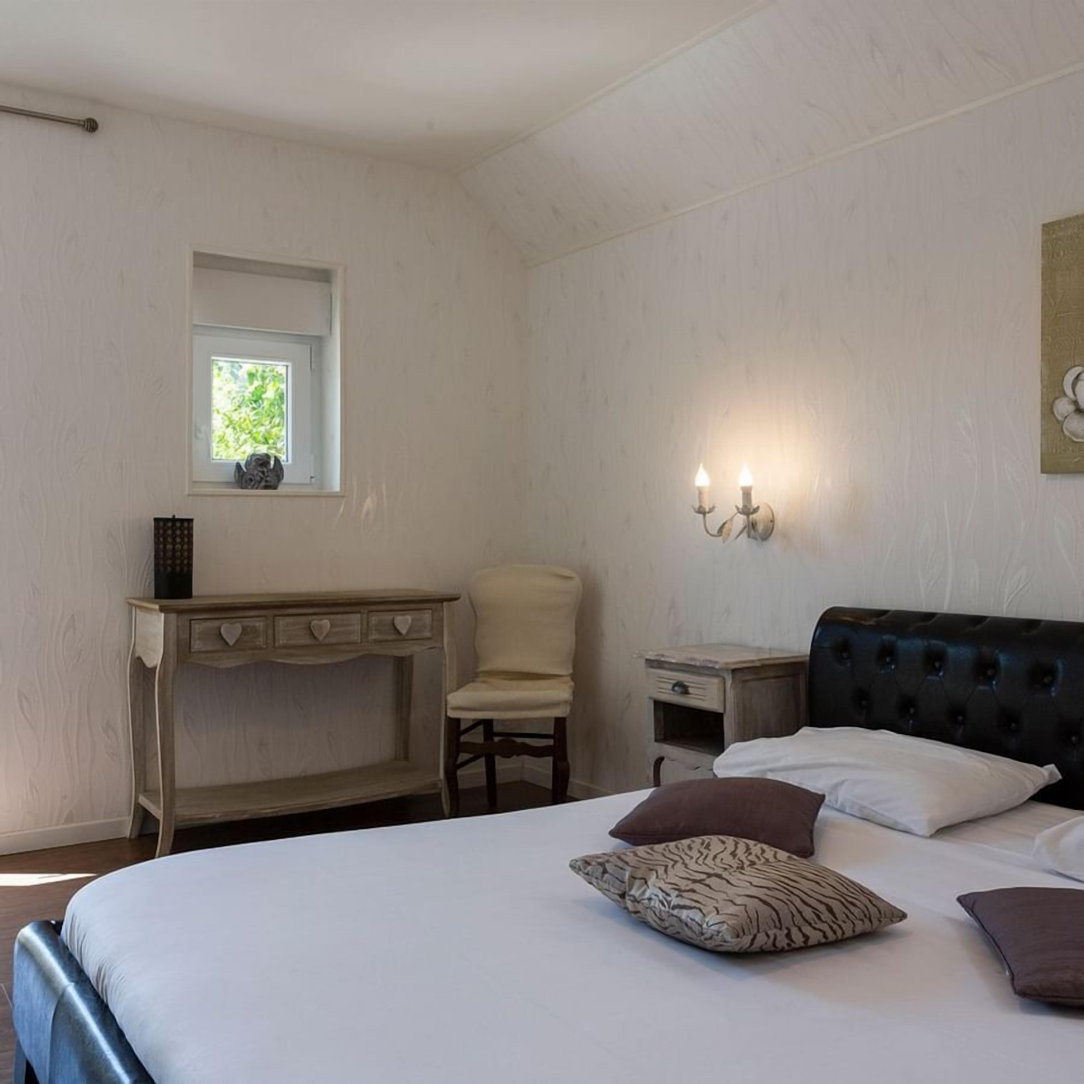 A bedroom with a bed, a desk, a chair, a nightstand, and a wall-mounted light fixture.