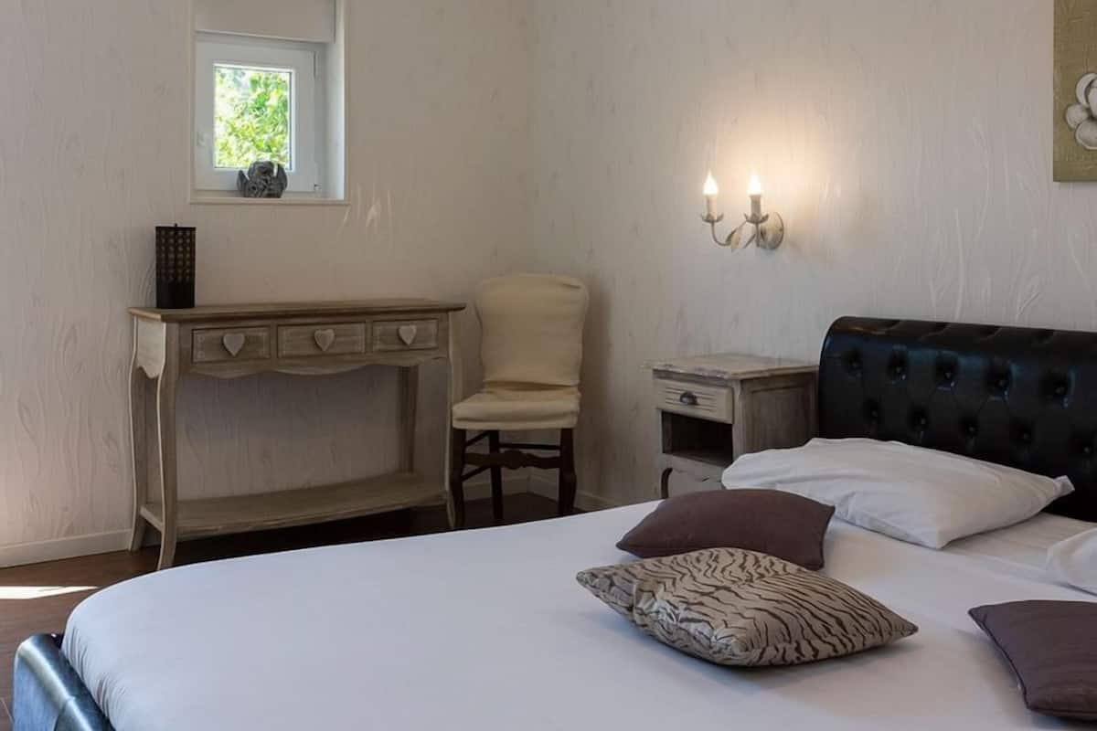 A bedroom with a bed, a desk, a chair, a nightstand, and a wall-mounted light fixture.