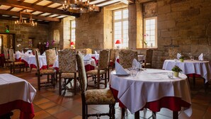Un restaurante con paredes de piedra, vigas de madera y candelabros. Las mesas están dispuestas con manteles blancos y servilletas rojas.