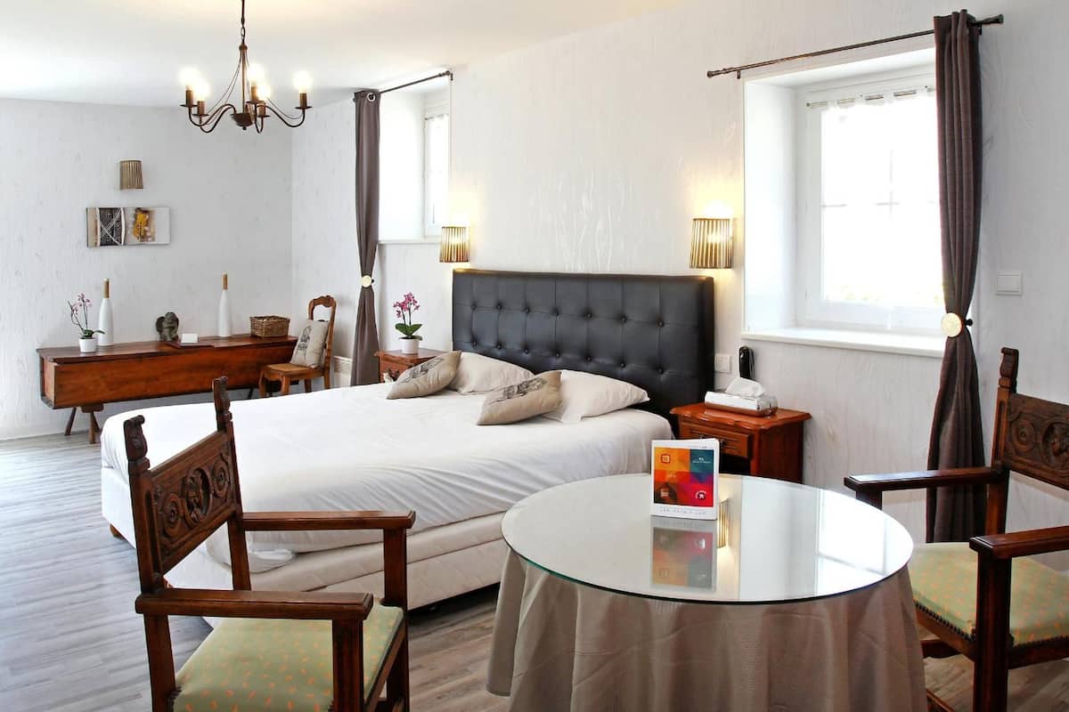 A bedroom with a large bed, a bedside table, a round glass table, two wooden chairs, a dresser, and a window with curtains.