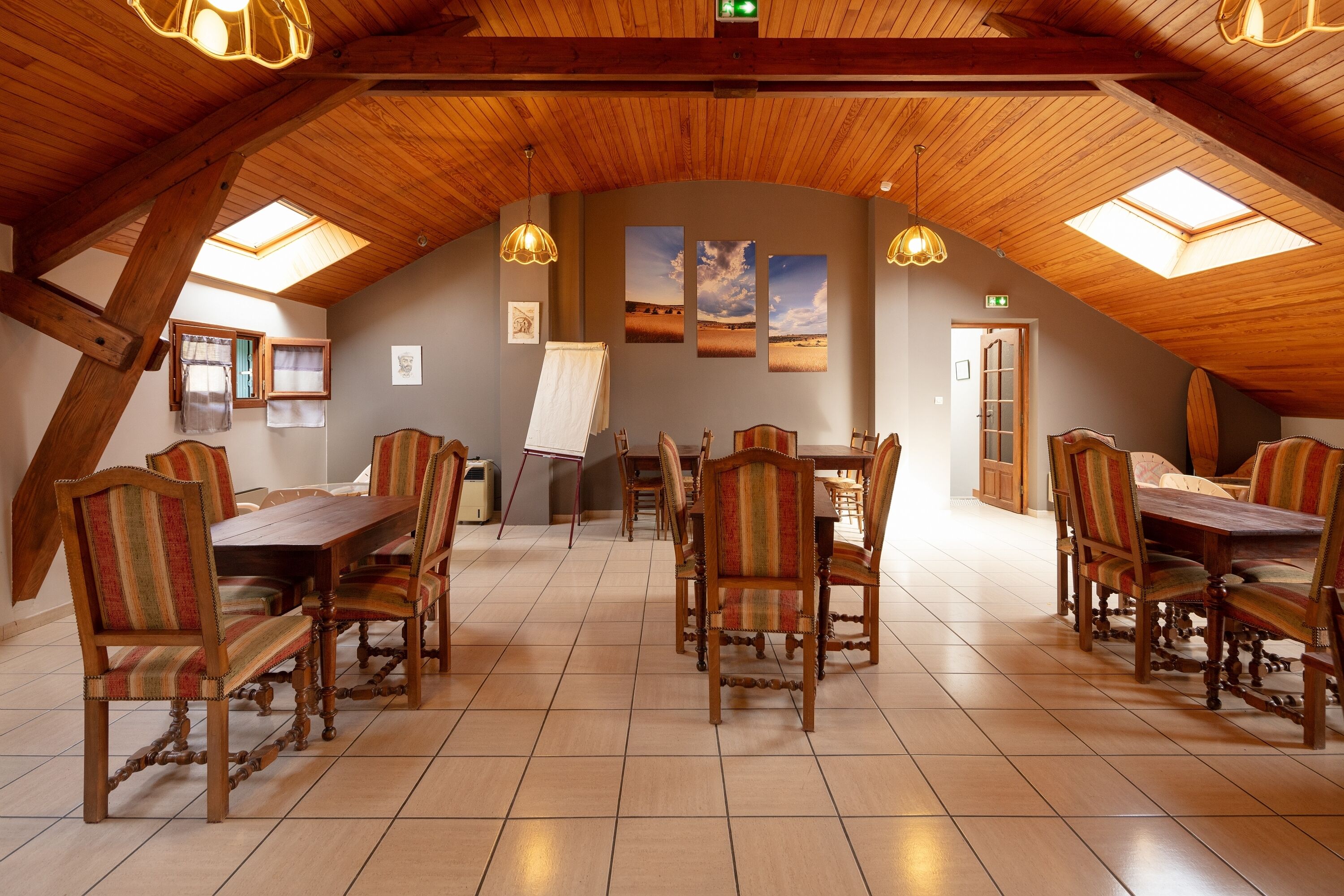 Un espace repas avec des tables et des chaises en bois, une lucarne et des tableaux encadrés accrochés au mur.