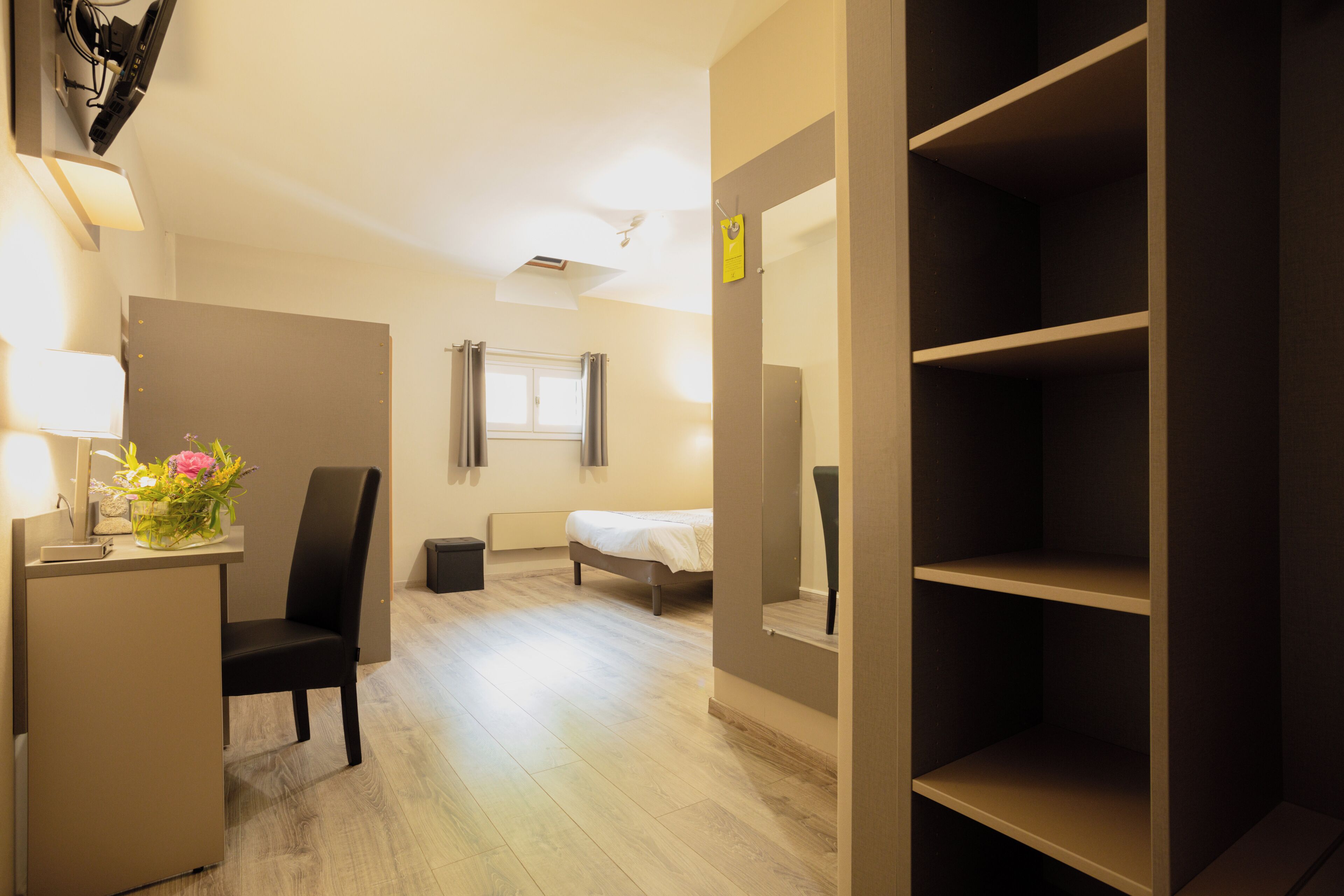 A hotel room with a bed, a desk with a lamp, a chair, and a shelf.