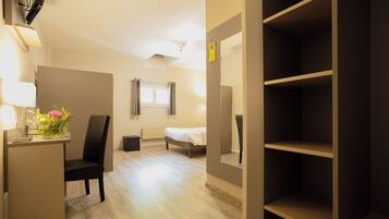 A hotel room with a bed, a desk with a lamp, a chair, and a shelf.