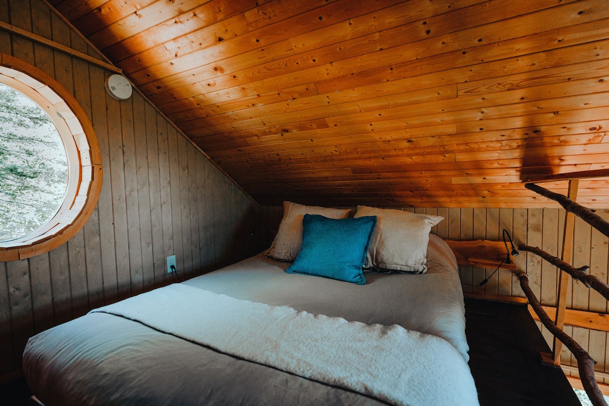 A wooden cabin bedroom with a bed, pillows, a window, and a wooden ladder.