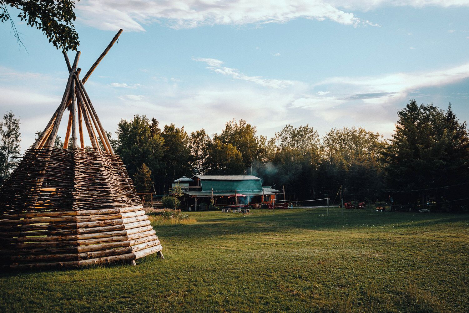 Un campo cubierto de hierba con una tienda tipi de madera y un edificio con tejado verde.