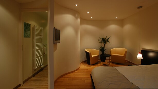 A modern hotel room with a curved wooden floor, a bed, two armchairs, a small table, and a potted plant. - Hôtel Saint-Pierre (Villedieu-les-Poêles-Rouffigny)
