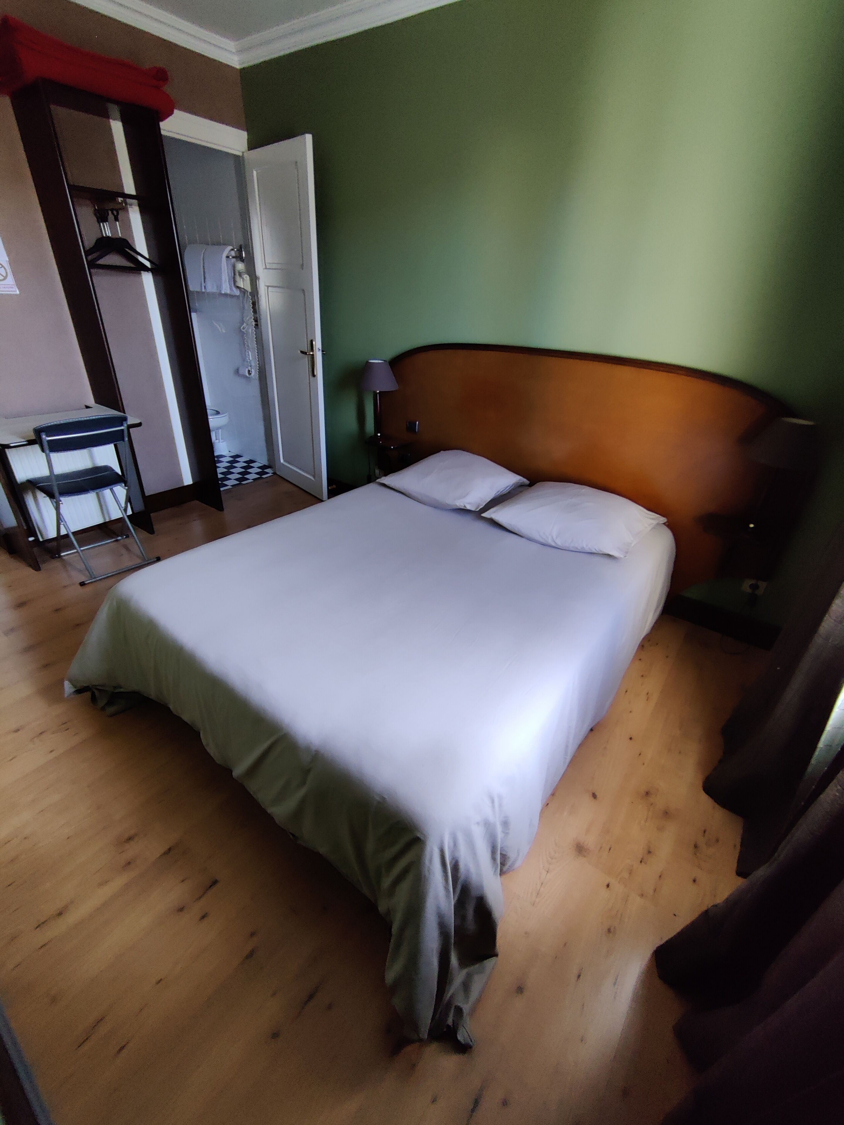 A hotel room with a bed, a nightstand, a lamp, a wooden floor, and a bathroom visible through an open door.