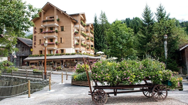 Un hotel de madera tradicional con balcones decorados con flores, rodeado de exuberante vegetación y un carro rústico lleno de flores.