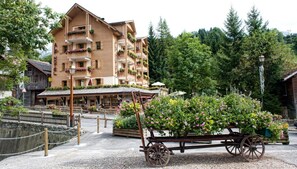 Un hotel de madera tradicional con balcones decorados con flores, rodeado de exuberante vegetación y un carro rústico lleno de flores.