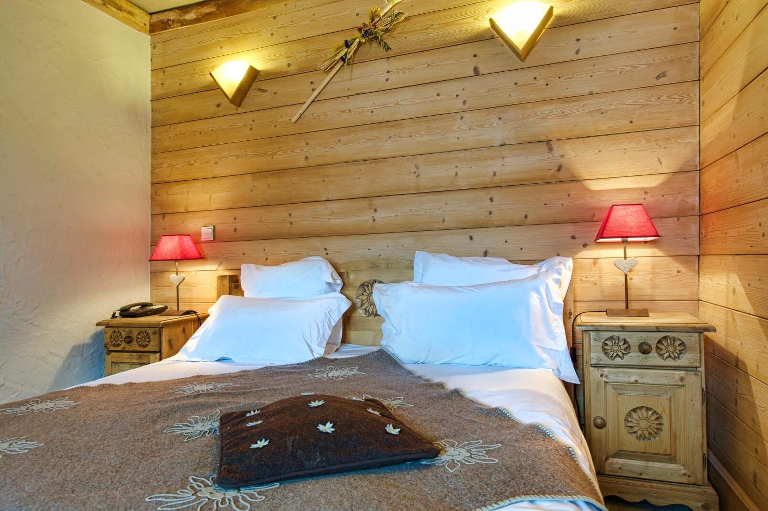 Ein schlichtes Schlafzimmer mit Holzwänden, ausgestattet mit zwei Betten, jeweils mit weißen Laken und einem braunen Zierkissen. Zwei Nachttische mit Lampen vervollständigen das Bild, eine rot und eine weiß.