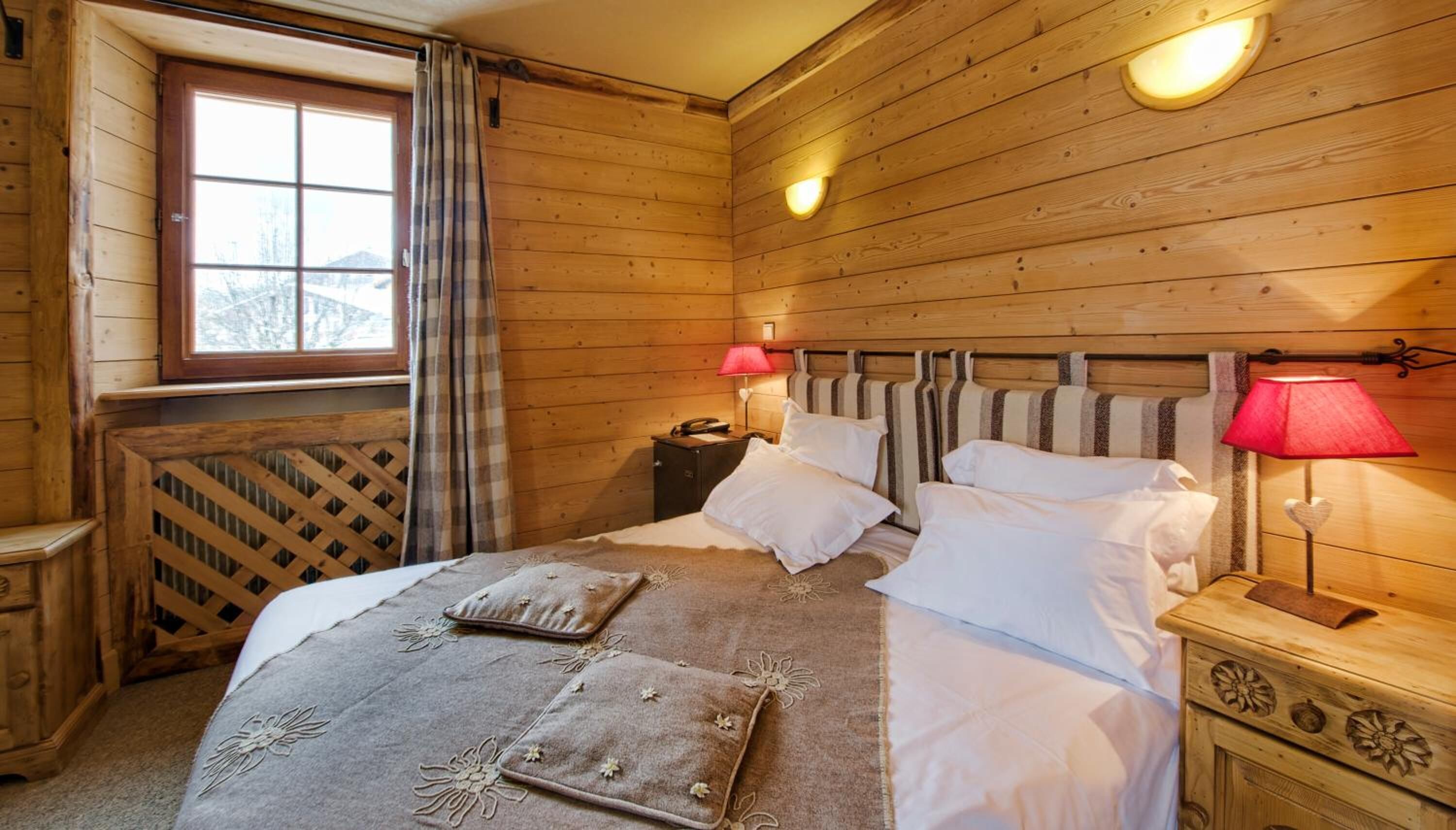 Una habitación de madera con una cama, dos mesitas de noche, una ventana con cortinas y un radiador.