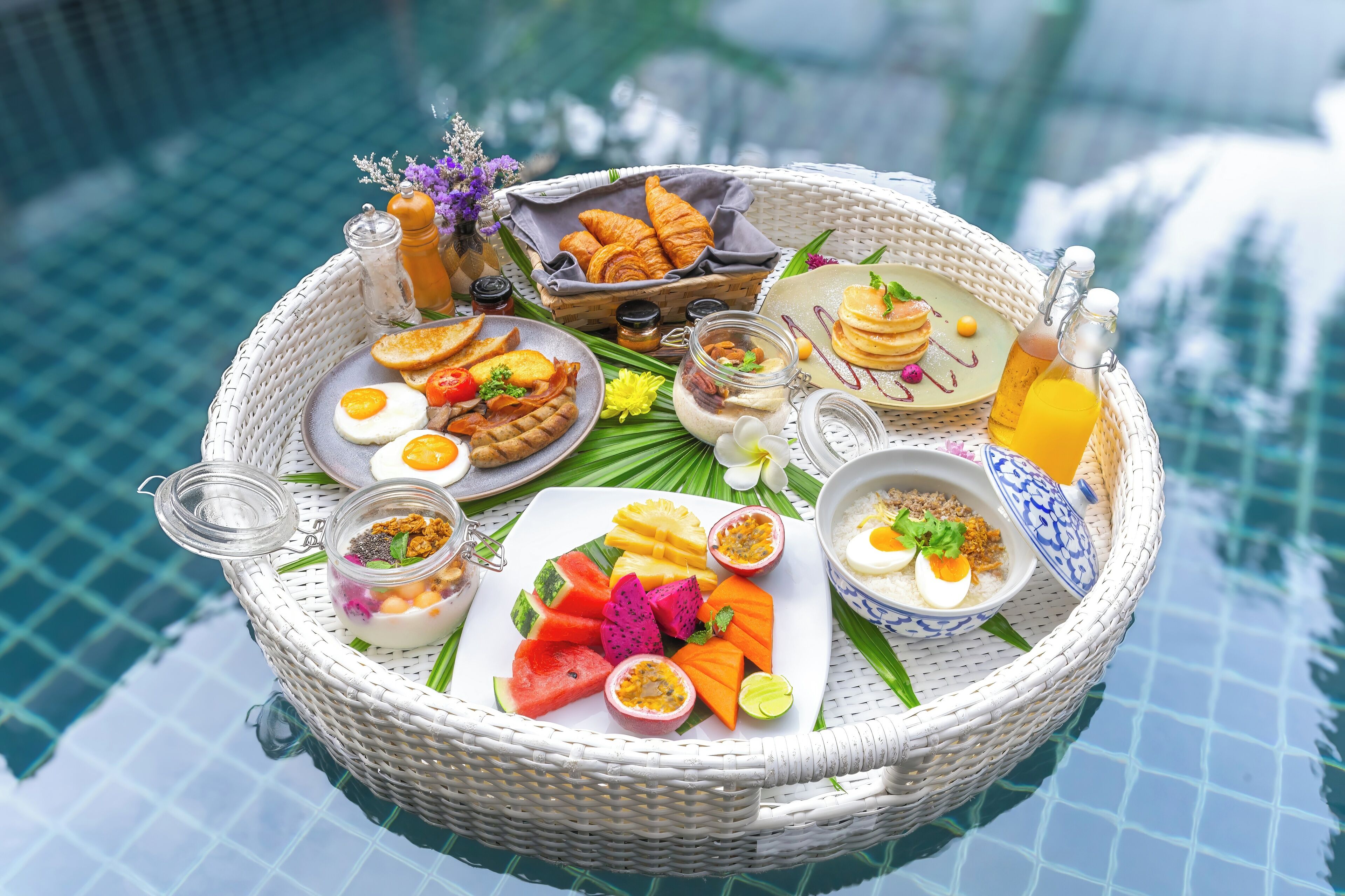 A wicker tray with breakfast items including eggs, pancakes, and fruit by a pool.