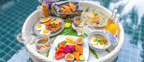 A wicker tray with breakfast items including eggs, pancakes, and fruit by a pool.